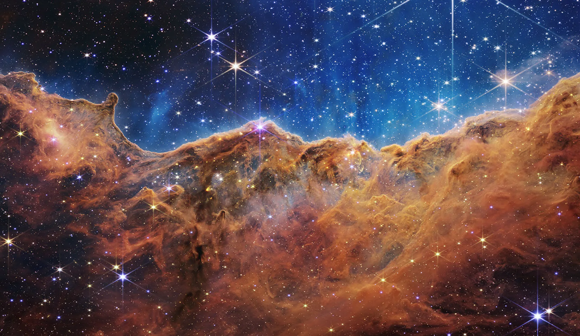 Webb telescope view of the Cosmic Cliffs in the Carina Nebula, with orange clouds beneath a blue field of stars.